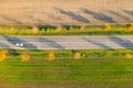 Road between green field and cultivated ground with yellow trees at sunset in autumn with car. Aerial view on asphalt speedway.