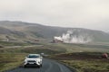 Road near the Krafla Geothermal Power Plant, as cars drive by. Steam rising from the plant
