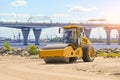 A road roller is at work on sandy terrain beside a river, with a large bridge and industrial cranes visible in the distance under Royalty Free Stock Photo
