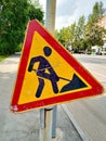 Road sign `Construction works`. The symbol is old, scuffed and dirty. Paint yellow and red. The black man is digging.