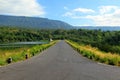 The road stretches along a beautiful river or lake, with mountains, blue sky, white cloud and green forests in the foreground