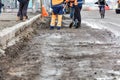 Road workers in orange and blue reflective overalls shovel debris into a pile on a road section being repaired