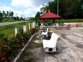 Roadside stone chairs with a red roof pavilion, red flowers and green leaves along the fence. Little garden at road side.