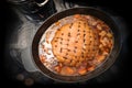 Roast pork with rind in diamond shapes and larded with cloves in a casserole with vegetables and broth on a black stove top,