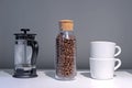 Roasted coffe grains in a glass jar, French press and two white coffe cups