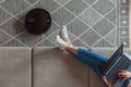 Robot vacuum cleaner cleaning carpet, woman using laptop sitting on sofa at home Royalty Free Stock Photo