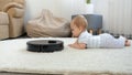 Robot vacuum cleaner rides to cute baby boy lying on carpet in living room. Concept of hygiene, household gadgets and Royalty Free Stock Photo