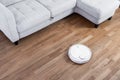 Robotic vacuum cleaner runs near sofa on laminate floor. Robot controlled by voice commands to direct cleaning. Modern smart