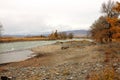 A rocky bank of a small river with yellowed trees at the beginning of a valley at the foot of high mountains