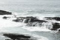 a rocky coastline with light blue, rough waves crashing against black, jagged rocks under an overcast light gray sky Royalty Free Stock Photo