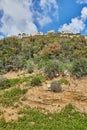 Rocky hill with a dirt road in front under a blue sky with white clouds and a modern villa on top