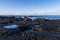 Rocky volcanic shoreline in Hawaii. Low tide; pools of water in rock cavities. Waves, ocean blue sky and clouds in background.