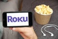 Roku logo on the mobile phone screen with popcorn box and Apple earpods on the background. Leisure time at home concept