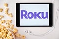 Roku logo on the tablet screen laying on the white table with scattered popcorn and Apple earphones. Spending free time