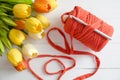 Roll of cotton yarn and a crochet hook with a bouquet of orange and yellow tulips on white wooden background.
