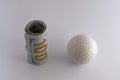 A roll of one hundred US dollar bills tied with a rubber band next to the golf ball on a white background. The concept of sports