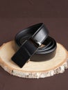 Rolled-up black fashionable men`s leather belt with automatic buckle on wood slice on brown background. Male accessory. Front vie Royalty Free Stock Photo