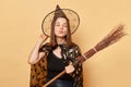Romantic charming beautiful woman wearing witch costume and cone hat holding broomstick isolated over beige background sending air