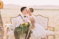 The romantic close-up portrait of the groom kissing the bride with the bouquet into the cheek in the sunny field. The