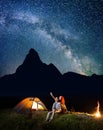 Romantic couple hikers looking at the shines starry sky at night. Happy pair sitting near camp and campfire