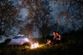 Couple tourists sitting at a campfire near tent, hugging each other under trees and night sky. Night camping Royalty Free Stock Photo