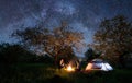 Couple tourists sitting at a campfire near tent under trees and night sky full of stars and milky way. Night camping