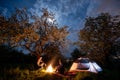 Couple tourists sitting at a campfire near tent under trees and night sky with the moon. Night camping Royalty Free Stock Photo
