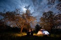 Couple tourists sitting at a campfire near tent under trees and night sky with the moon. Night camping Royalty Free Stock Photo