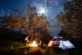 Romantic couple tourists sitting at a campfire near tent under trees and night sky with the moon Royalty Free Stock Photo
