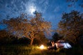 Romantic couple tourists sitting at a campfire near tent under trees and night sky with the moon Royalty Free Stock Photo