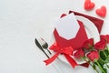 Romantic creative table setting. Empty white plate and red roses, candles, knife, fork and decorative silk hearts on white