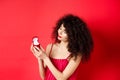 Romantic curly-haired woman in red dress, looking happy at engagement ring, become a bride, standing on studio