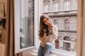 Romantic dark-haired girl spending time at home and drinking latte on sill. Portrait of dreamy caucasian young woman Royalty Free Stock Photo