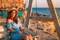 Romantic evening at sunset by the sea, a young woman in a dress with a bouquet of flowers is sitting on a wooden swing holding her Royalty Free Stock Photo