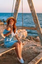 Romantic evening at sunset by the sea, a young woman in a dress with a bouquet of flowers is sitting on a wooden swing holding her Royalty Free Stock Photo