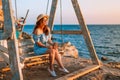 Romantic evening at sunset by the sea, a young woman in a dress with a bouquet of flowers is sitting on a wooden swing holding her Royalty Free Stock Photo