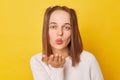 Romantic funky teenage girl with ponytails in jumper posing isolated over yellow background sending air kissing flirting with her