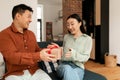 Romantic gift. Asian man giving wrapped box to his wife, celebrating anniversary or birthday, sitting at home
