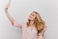 Romantic girl with shy smile making selfie while playing with blonde hair. Indoor portrait of charming young woman in Royalty Free Stock Photo