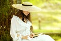 Romantic Girl in White Dress and Straw Sun Hat reading Book sitting under Tree. Young Woman studying in Spring Park. Old Fashioned Royalty Free Stock Photo