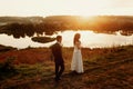 Romantic landscape, newlywed couple posing at sunset field near a lake, handsome groom walking towards gorgeous bride Royalty Free Stock Photo