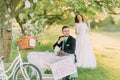 Romantic picknick in park. Playful bride at background and her stylish groom drinking tea on front