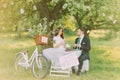 Romantic picknick under tree in park. Happy bride drinking tea with her lovely new husband. Decorated bicycle stands near Royalty Free Stock Photo