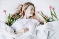 Romantic portrait of a girl in bed with natural flowers. Young woman with tulips in hair resting in bedroom in morning.