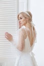 Romantic portrait of a woman near the window in a beautiful long white dress at home. The girl is blonde with blue eyes and Royalty Free Stock Photo
