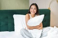 Romantic smiling asian girl lying in her bed, hugging pillow and gazing dreamy, lazy mornings in bedroom