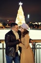 Romantic stylish couple in warm clothes embracing, looking at each other with smile, standing against ice rink with Royalty Free Stock Photo