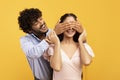 Romantic surprise concept. Portrait of happy indian guy covering his pretty girlfriend eyes from back, yellow background Royalty Free Stock Photo