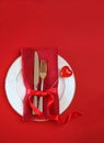 Valentines day table setting with white plate fork and knife shape heart on red tablecloth with ribbon bow background. Romantic Royalty Free Stock Photo