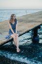 Romantic traveler woman in a blue dress is enjoying a beach holiday. A happy girl is sitting on the dock, looking into Royalty Free Stock Photo